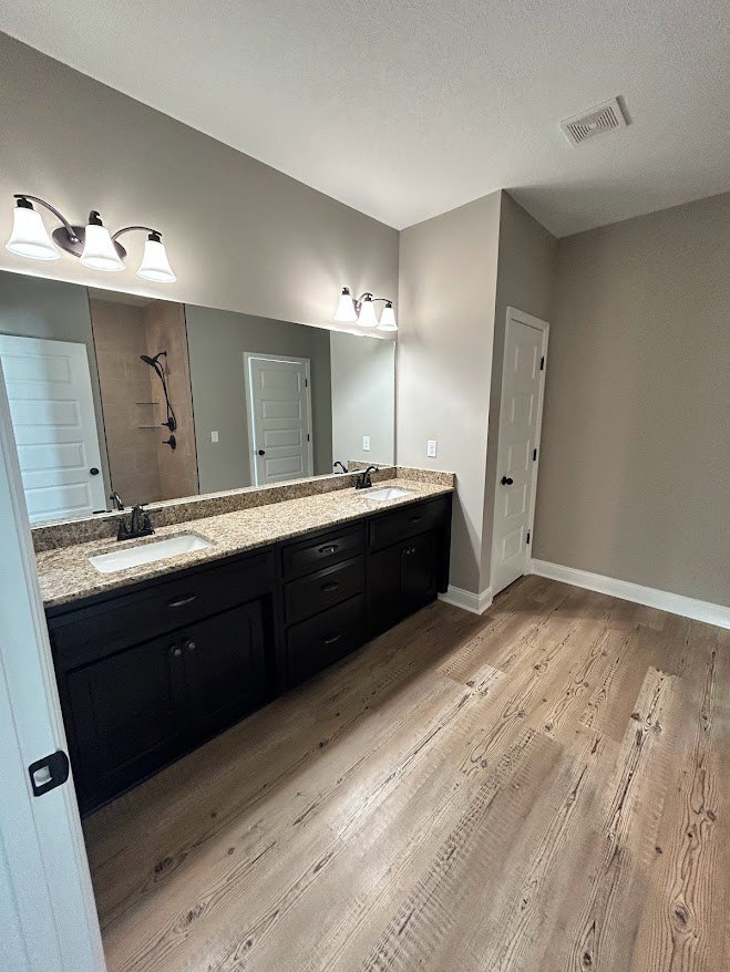 Bathroom featuring double sinks set in a stone countertop, large wall mirror above, white cabinetry below, and a three-light fixture mounted over the mirror; neutral tile flooring