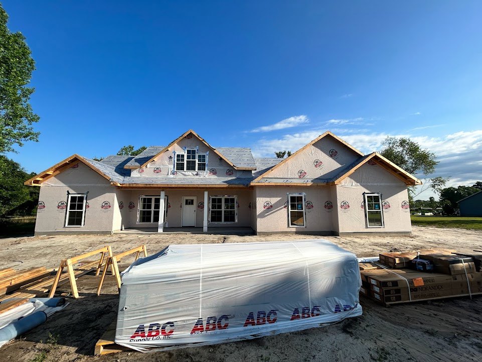 Partially built house with exposed framing, white-framed window, roof under construction, pallets and stacked boxes on ground, blue tarp covering nearby truck, clear sky overhead.