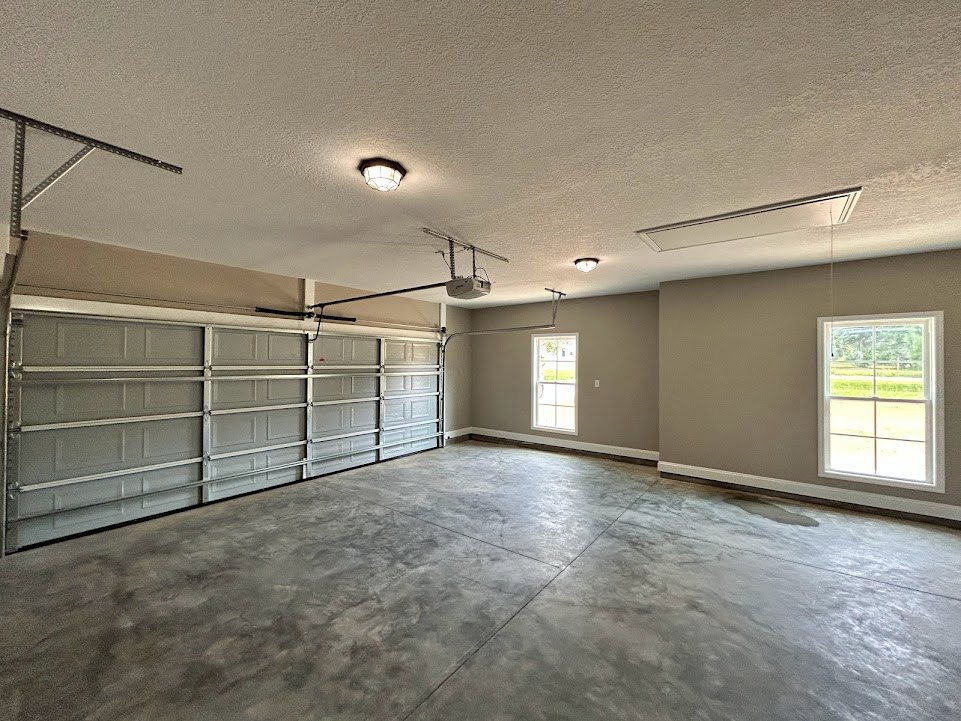 Spacious garage with smooth concrete floor, white plaster walls, large window framed in white overlooking grass and trees, sectional garage door, and clean white ceiling