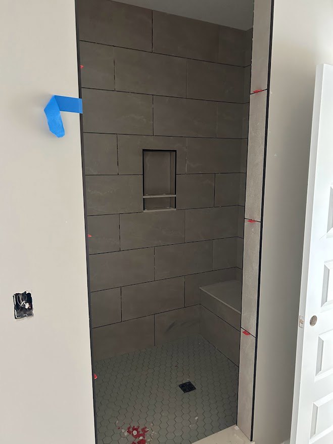 Bathroom with a tiled shower wall, built-in shelf, white and grey walls, shower floor drain, blue painter’s tape on a white surface, red and white paint marks on tile floor