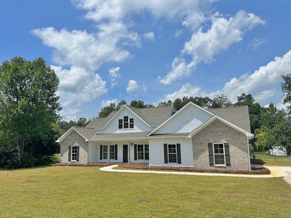 White house with black shutters, brick foundation, large front lawn, mature leafy trees in background, windows facing the yard
