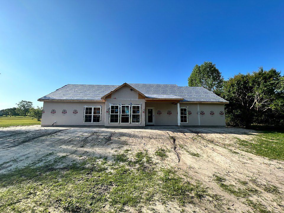 Two-story house with gray roof, white siding, large multi-pane windows, surrounded by dirt field and mature trees under clear blue sky