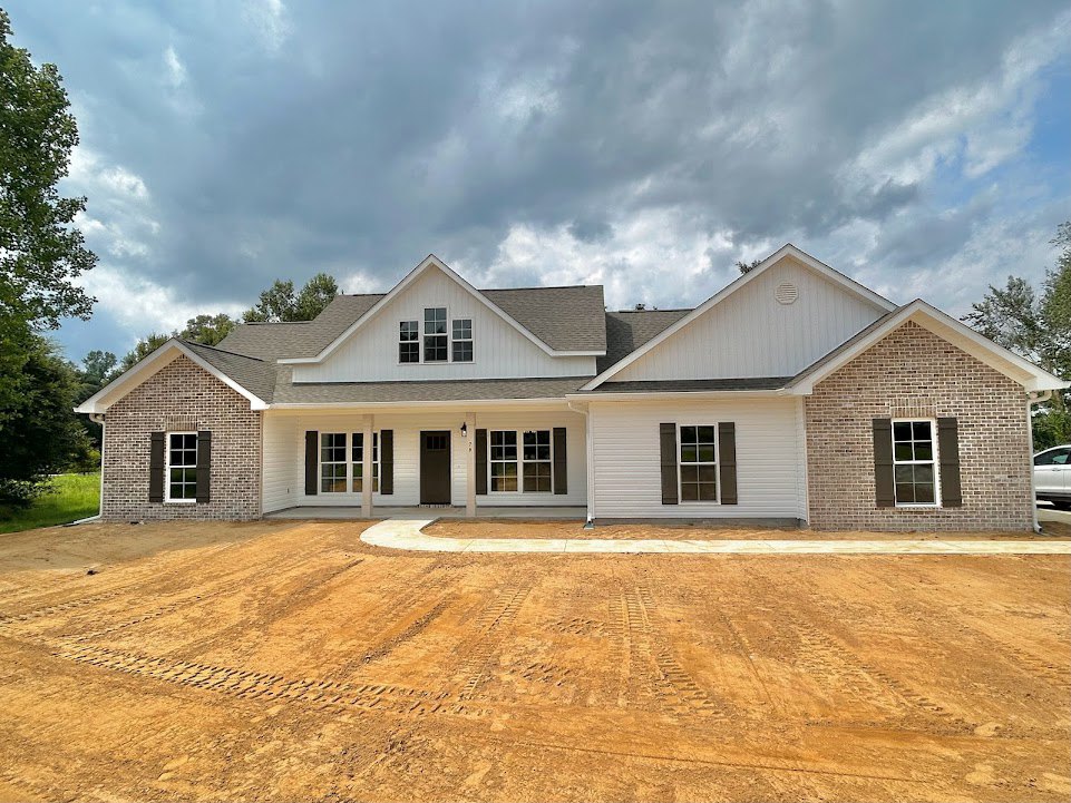 Grey-roofed house with white-framed windows, dirt driveway marked by tire tracks, cloudy sky overhead, surrounded by trees.