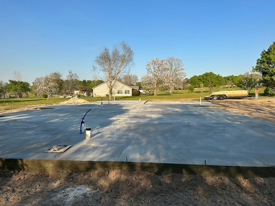 Concrete slab foundation surrounded by dirt and metal beam, with leafy trees and white flowering plants in the background.