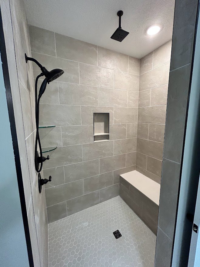 Walk-in shower with two matte black shower heads, white tile walls, glass shelf mounted on side wall, plaster ceiling above