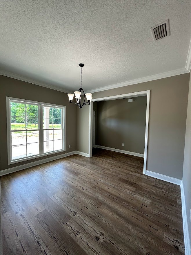 Wood flooring in a spacious room with a ceiling chandelier, white plaster walls, large window overlooking trees, wall vent, and door frame.