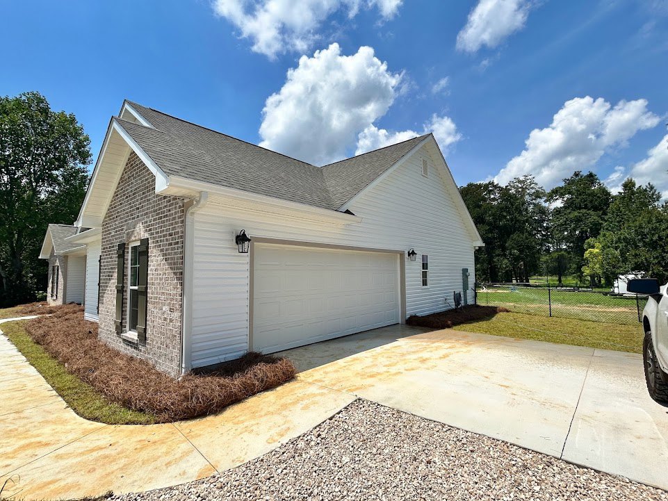 Two-story house with white garage door, brown mulch landscaping, concrete driveway, wooden fence, and large front windows under a cloudy sky