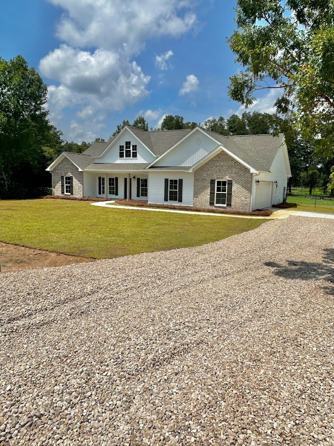White house with brick accents and brick wall, gravel driveway bordered by green lawn, mature trees casting shadows on grass, blue sky overhead