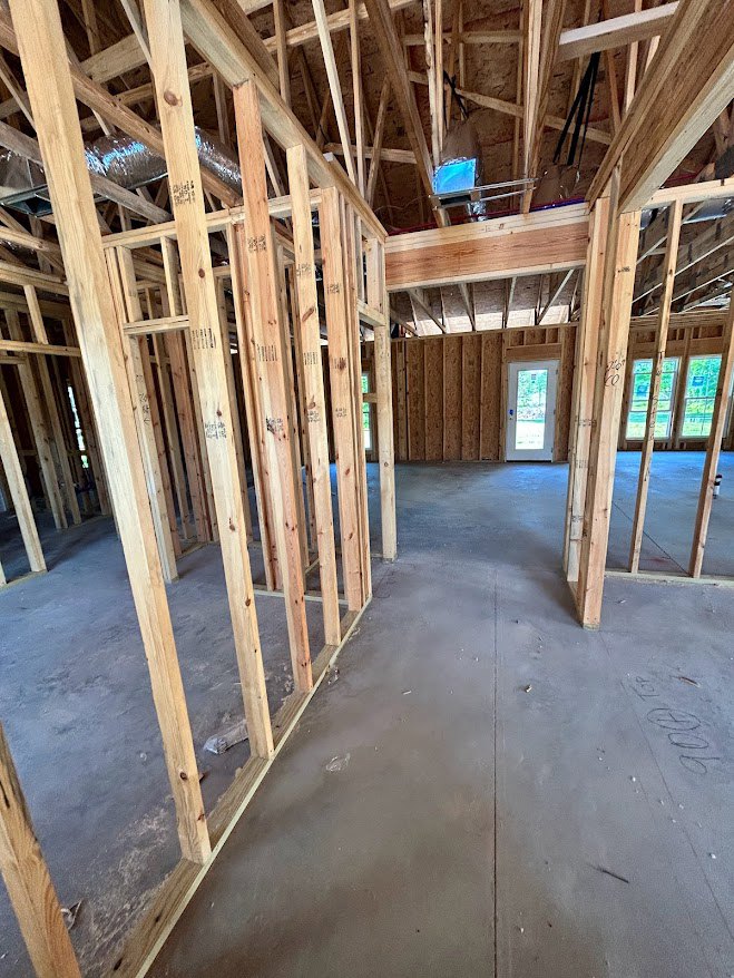 Exposed wood framing and beams inside a partially constructed home, concrete floor, glass door, window overlooking greenery, snow with writing visible outside