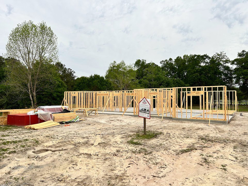 Wood-framed house under construction on dirt lot, surrounded by tall trees and cloudy sky, construction sign with house illustration in foreground