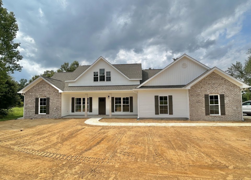 Two-story house with grey roof, white-framed windows, and dirt driveway with tire tracks, surrounded by green trees under a cloudy sky; Robert Frost Farm visible in the background.