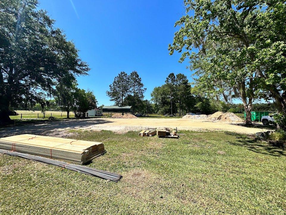 Grassy construction site with stacked wood planks, metal rods, and leafy tree in background