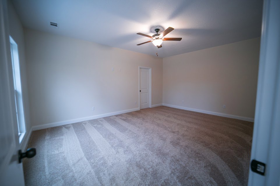 Carpeted bedroom with white walls, ceiling fan with light, and white door featuring a black knob