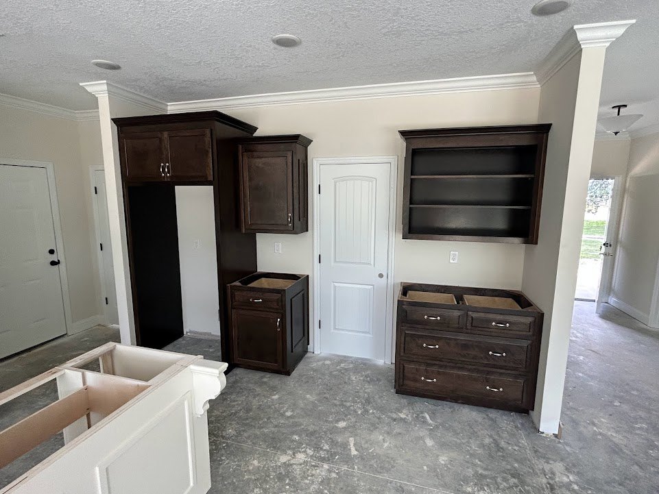 Kitchen featuring dark wood cabinets, white door with round knob, built-in wall shelf, black cabinet with drawer, white cabinet with multiple drawers, light flooring, and neutral
