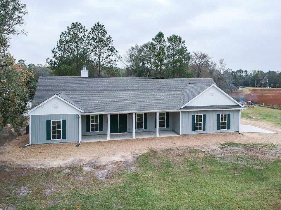 Two-story house with grey and white roof, white-framed windows, surrounded by mature trees, dirt path running alongside the exterior