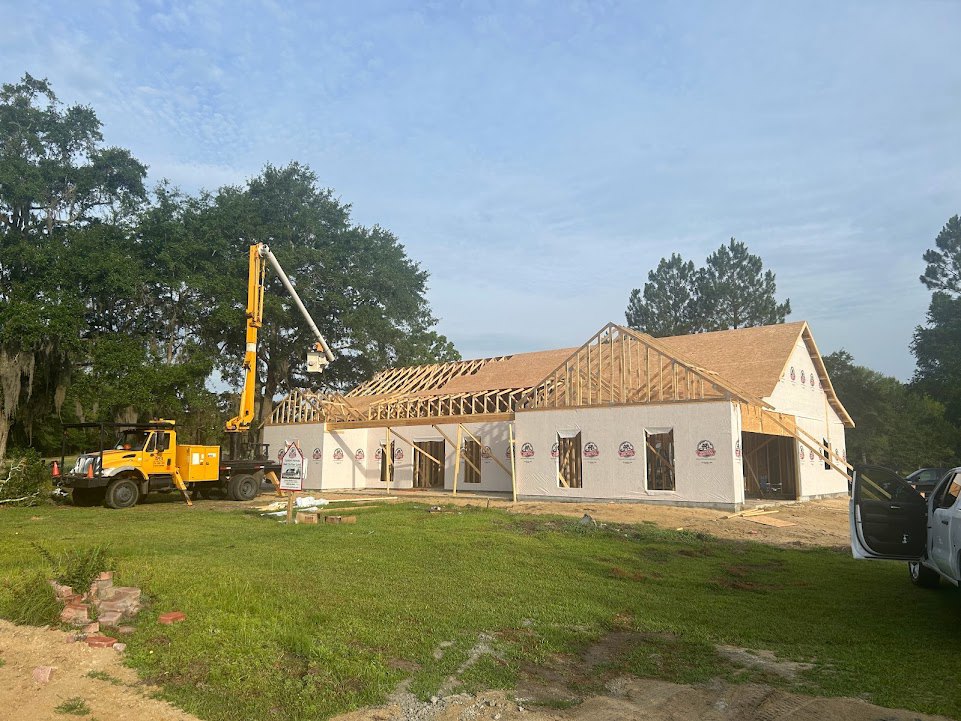 Wood-framed house under construction with exposed roof trusses, yellow crane truck parked on grassy lot, dirt patches and scattered building materials visible