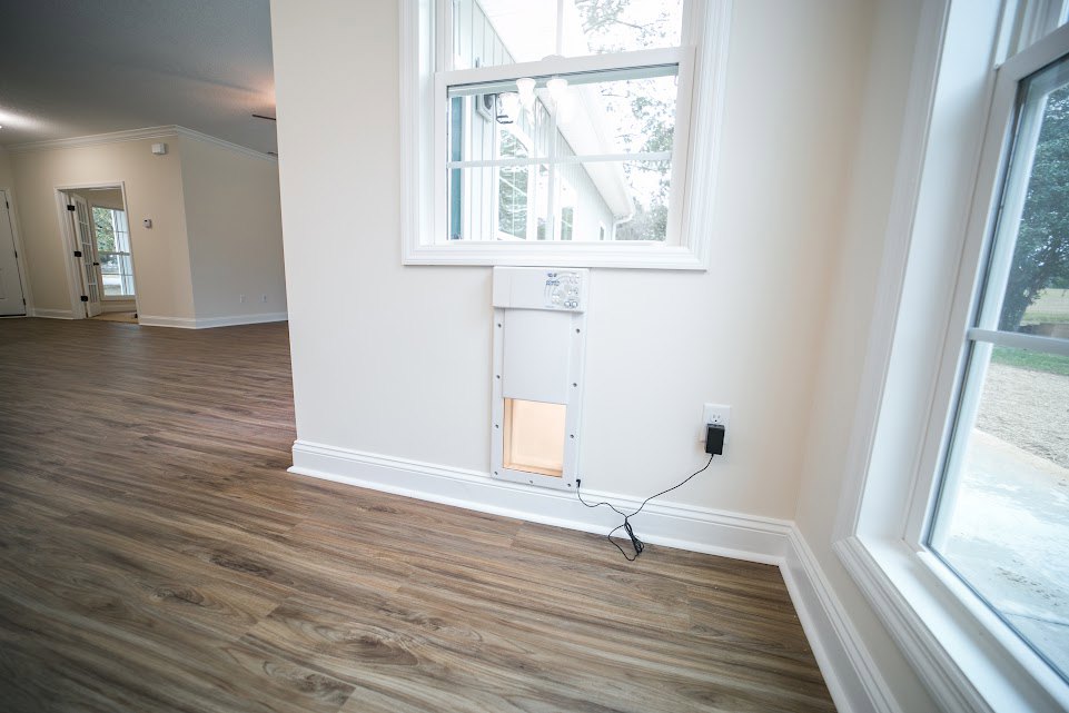 White plaster wall, wide plank wood flooring, rectangular window with white trim, ceiling light fixture, black electrical cord emerging from wall.