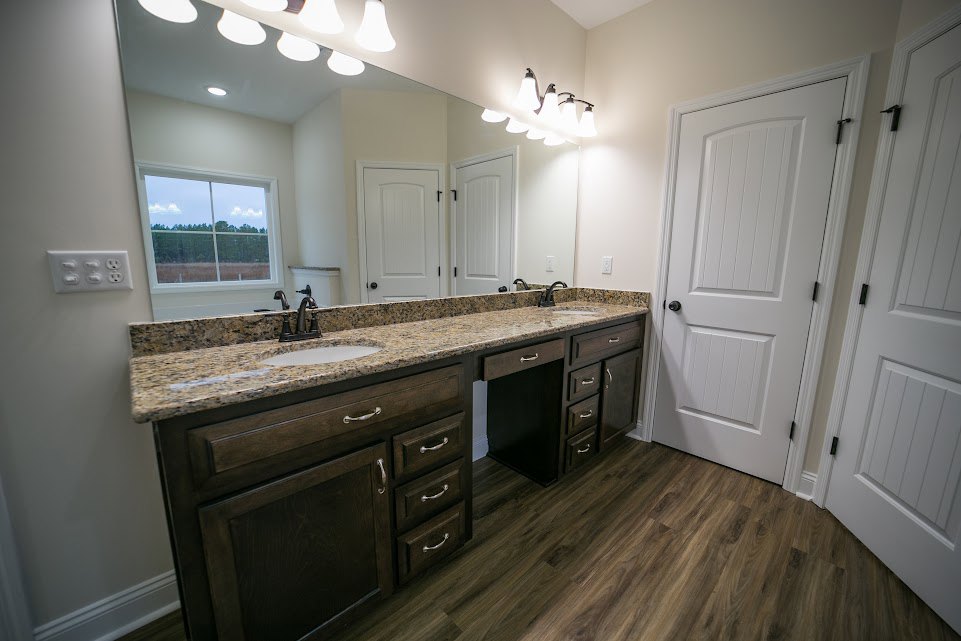 Bathroom with double vanity featuring white sinks and stone countertop, wood plank flooring, white door with black hardware, window overlooking trees, wall-mounted row of lights