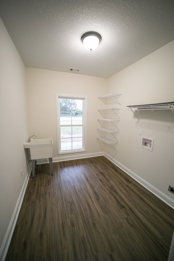 Wood floor room with white sink, wall-mounted white shelves, plaster walls, ceiling fixture, and window overlooking grassy field.