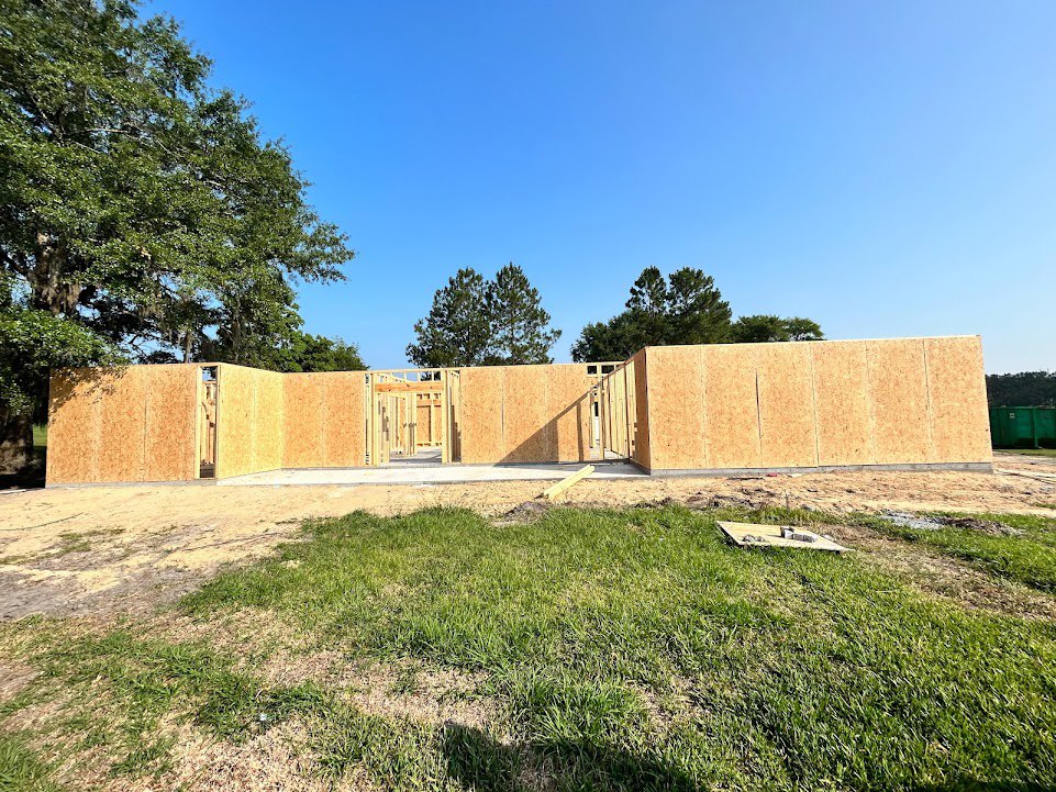 Wood-framed house under construction surrounded by green grass and mature trees, blue sky overhead