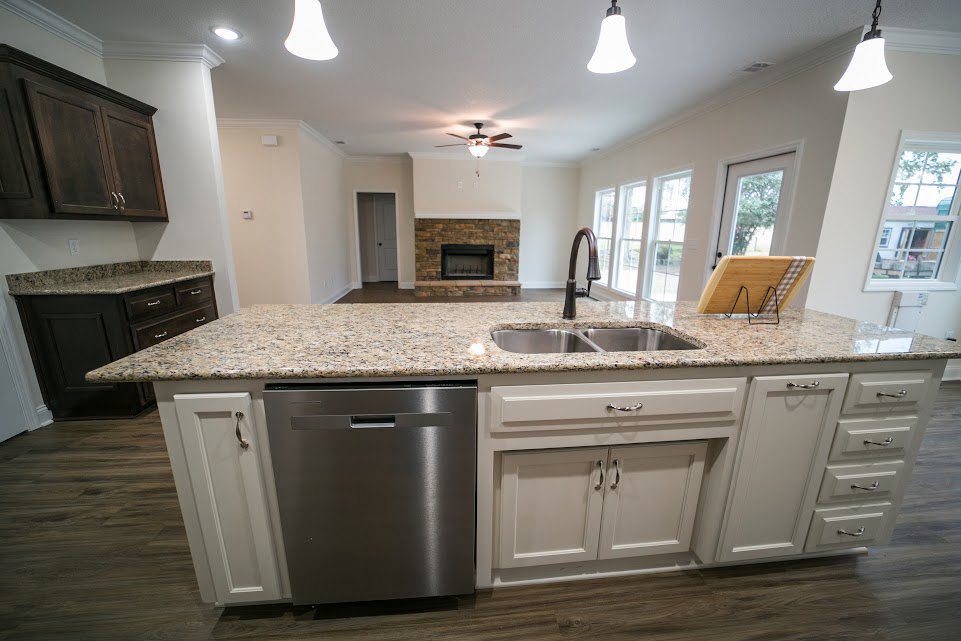 Granite countertop kitchen with stainless steel sink, tile backsplash, wood cabinetry, built-in dishwasher, and brick fireplace