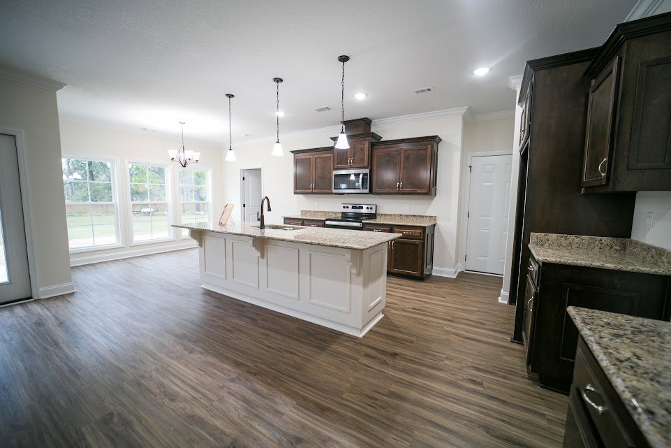 Open kitchen and dining area featuring wood flooring, central kitchen island with marble countertop, white cabinetry, stainless steel sink, large windows, dining table, and white