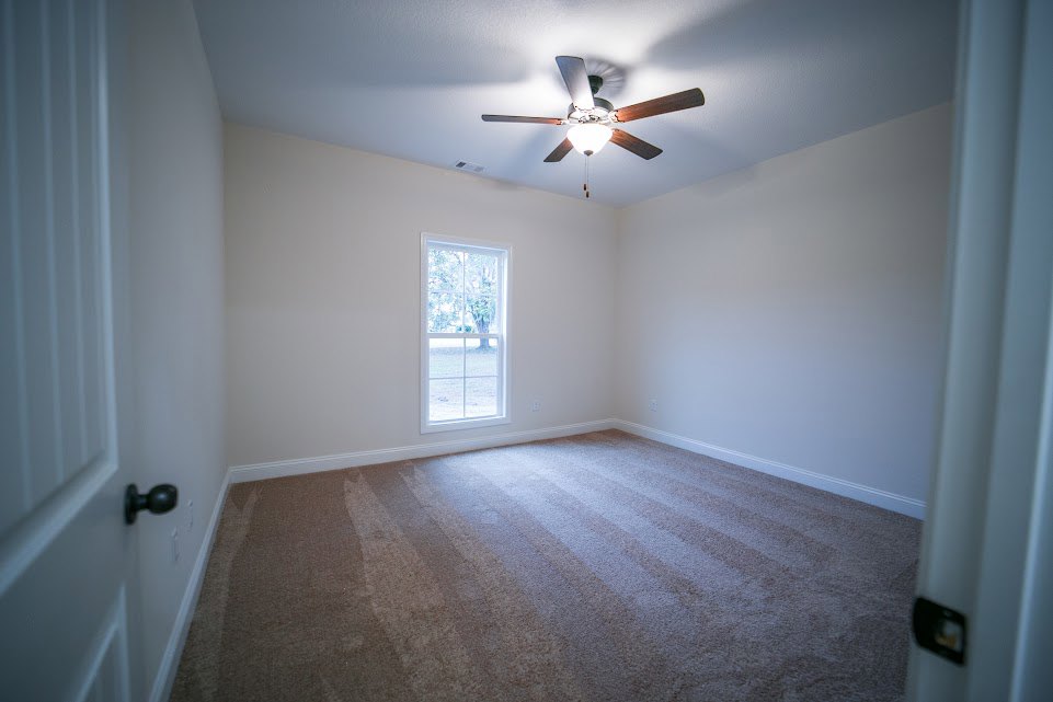 Carpeted bedroom with white walls, ceiling fan with light fixture, large window overlooking trees, white trim and molding