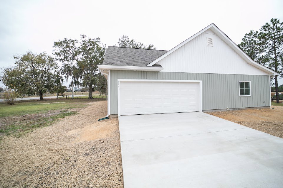 White garage door beneath a gray roof, concrete driveway leading to the entrance, light-colored siding, leafy tree beside the house, blue sky overhead