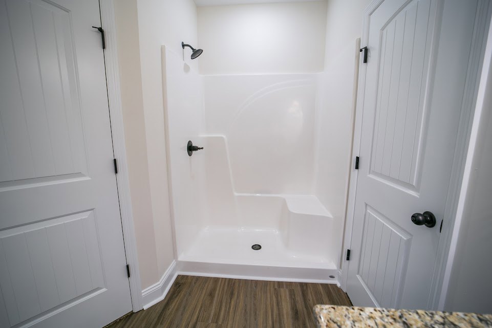 Walk-in shower with glass enclosure, white tile walls, wood-look flooring, white door featuring black handle and knob