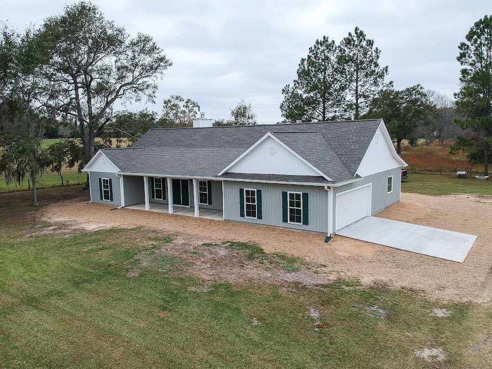 Two-story home with grey shingle roof, white siding, concrete driveway, green lawn, large tree in background, and multiple windows.