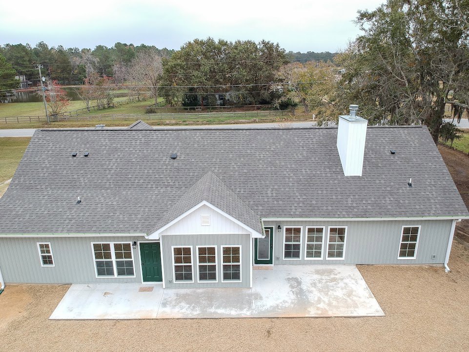 Grey siding house with green front door, white framed windows, paved driveway, mature trees, and a white rooftop utility box.