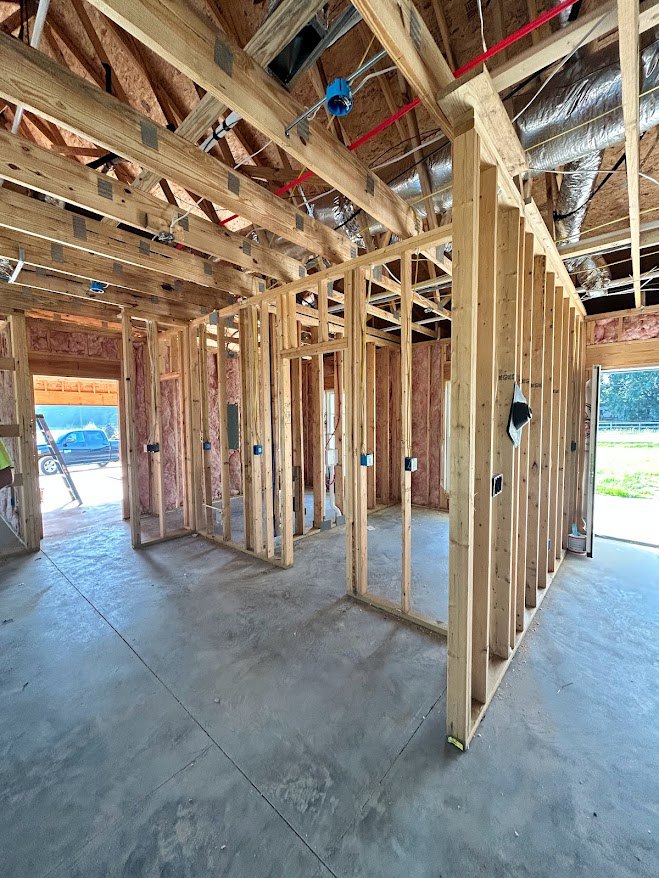 Exposed wood framing and ceiling beams with unfinished concrete floor, window opening facing fenced yard, visible plumbing pipes and insulation