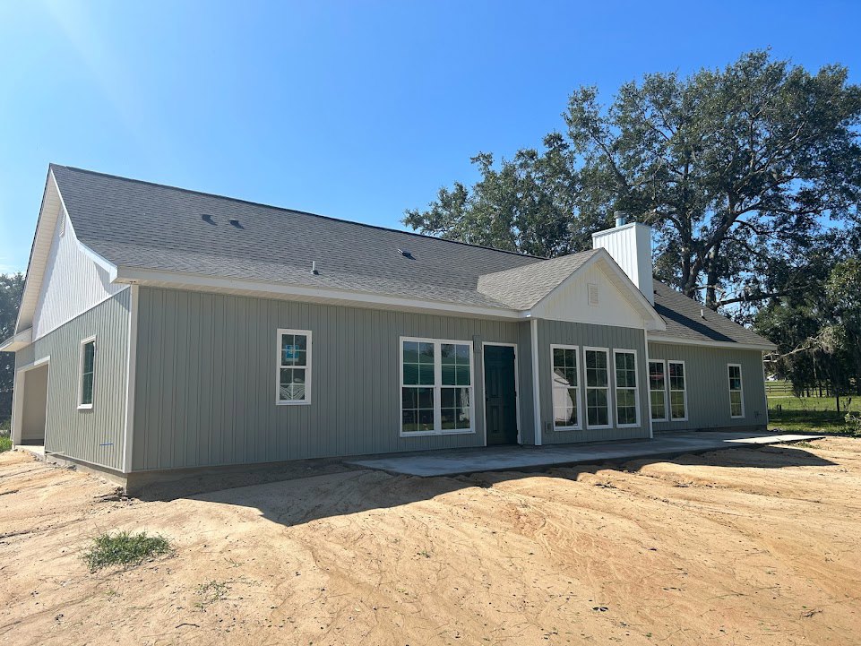Grey siding house under construction with white door and multiple windows, dirt patch in front, surrounded by trees and unfinished landscaping.