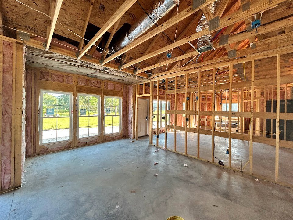 Open room with exposed wood ceiling beams, large windows overlooking grassy field, concrete floor, visible wood framing, metal pipe along ceiling, window with sign attached