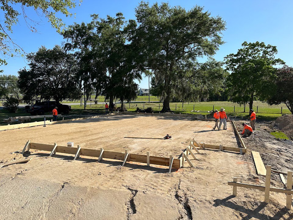 Crew in orange jackets working beside wooden beams and a large excavation pit, surrounded by dirt, trees, and open sky on a custom home construction site