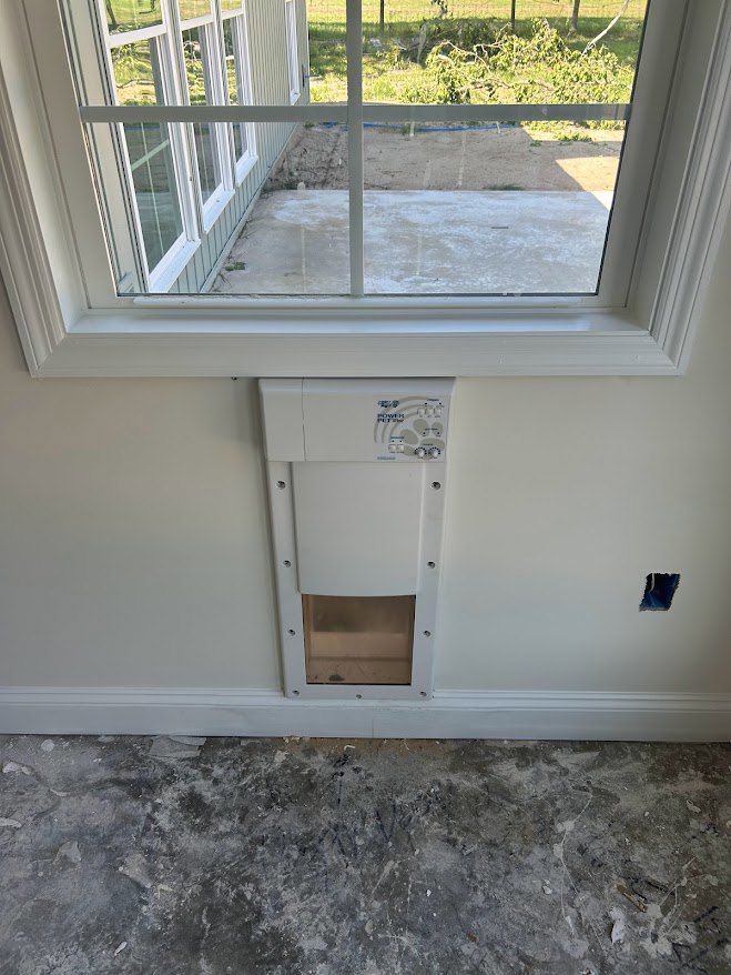 Large window overlooking a grassy yard, grey concrete floor with black bird prints, white rectangular vent with holes, white walls, natural daylight filling the room