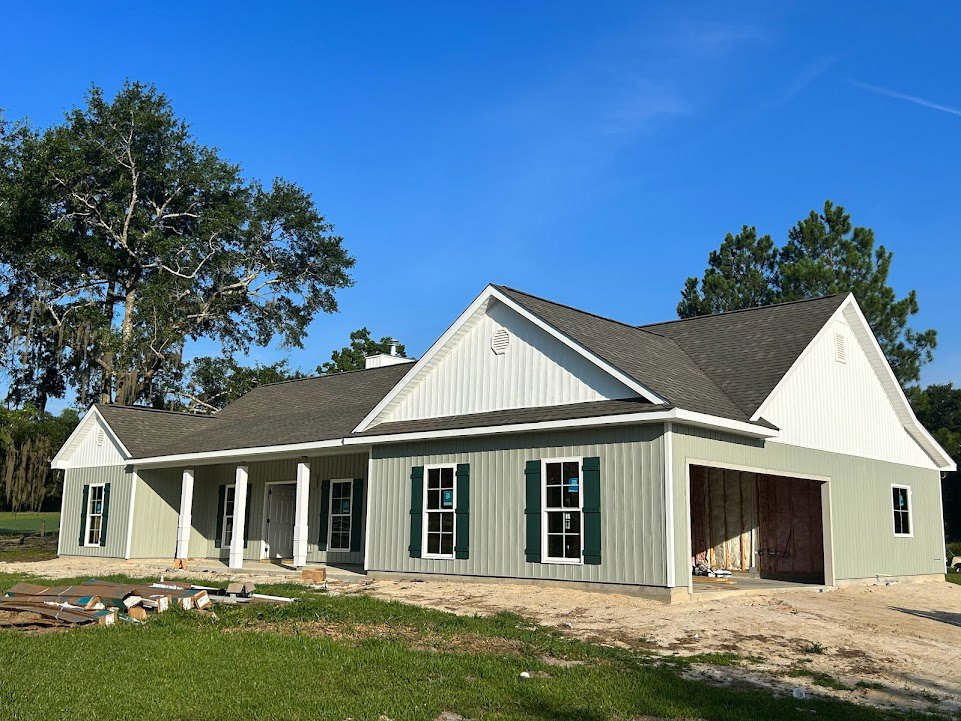 Partially built home with exposed framing, white-framed windows, and garage door; surrounding trees and a small white cottage visible in the background