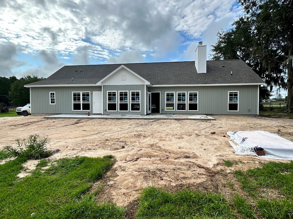 Partially built house with exposed framing, surrounded by grass and mature trees, cloudy sky overhead, dirt patch in front yard, white tarp covering construction materials