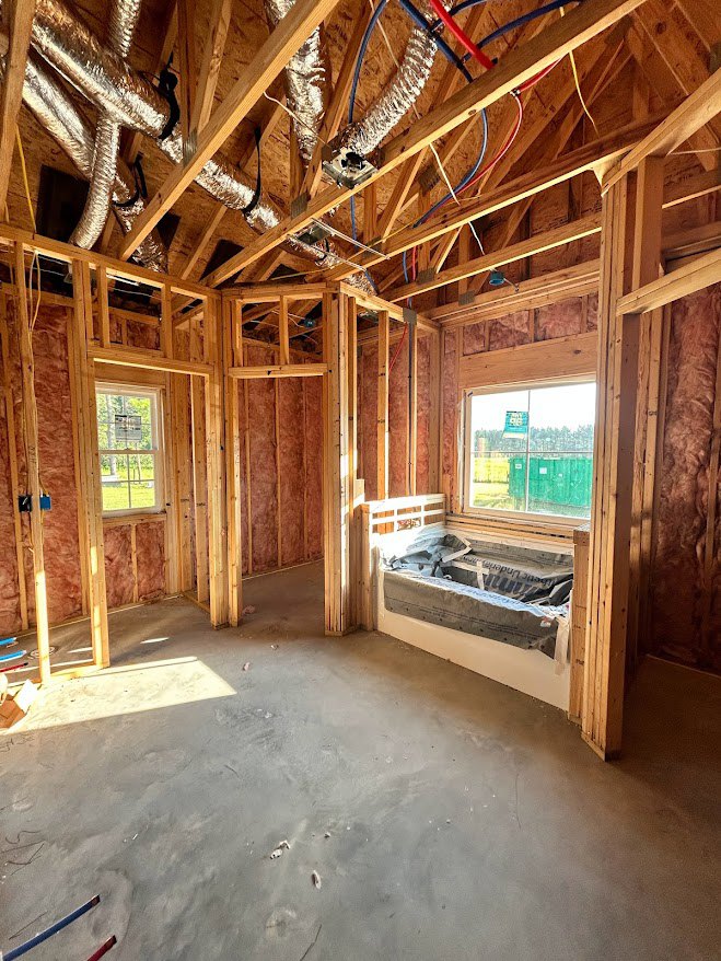 Freestanding bathtub beside large window, concrete floor, wood ceiling beams, unfinished walls with insulation, built-in bench along one wall