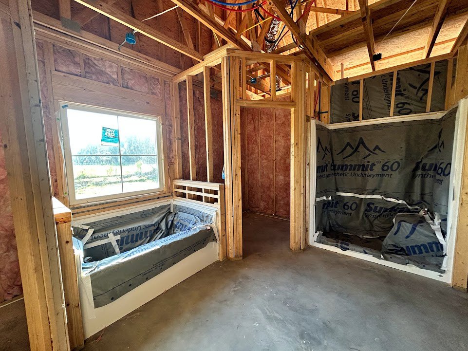Freestanding bathtub positioned beneath a large window, unfinished walls with exposed lumber and plastic sheeting, construction materials visible on floor