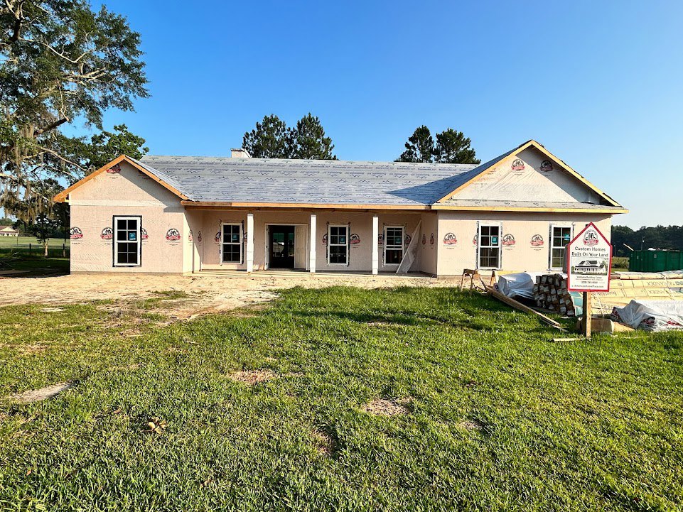 White farmhouse-style home with dark roof, glass-paneled front door, grassy yard with patches of dirt, tree branches near roof, and a sign displaying a house illustration in front.