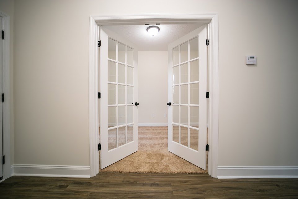 White double doors with glass panes, surrounded by white molding and plaster walls, opening into a bright interior room with wood flooring