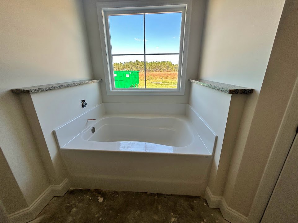 Freestanding white bathtub with chrome faucet beside a large window, natural light illuminating tiled bathroom walls, green dumpster visible outside