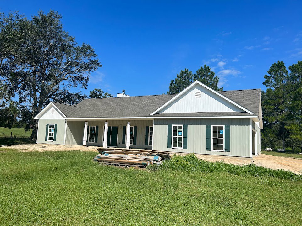 White house with covered porch, large green yard, mature tree, boats and wood pile near the front, blue sticker on window