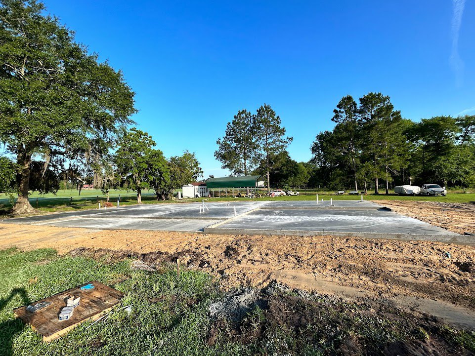 Concrete foundation slab surrounded by dirt and grass, several upright poles in the middle, leafy trees and blue sky in the background