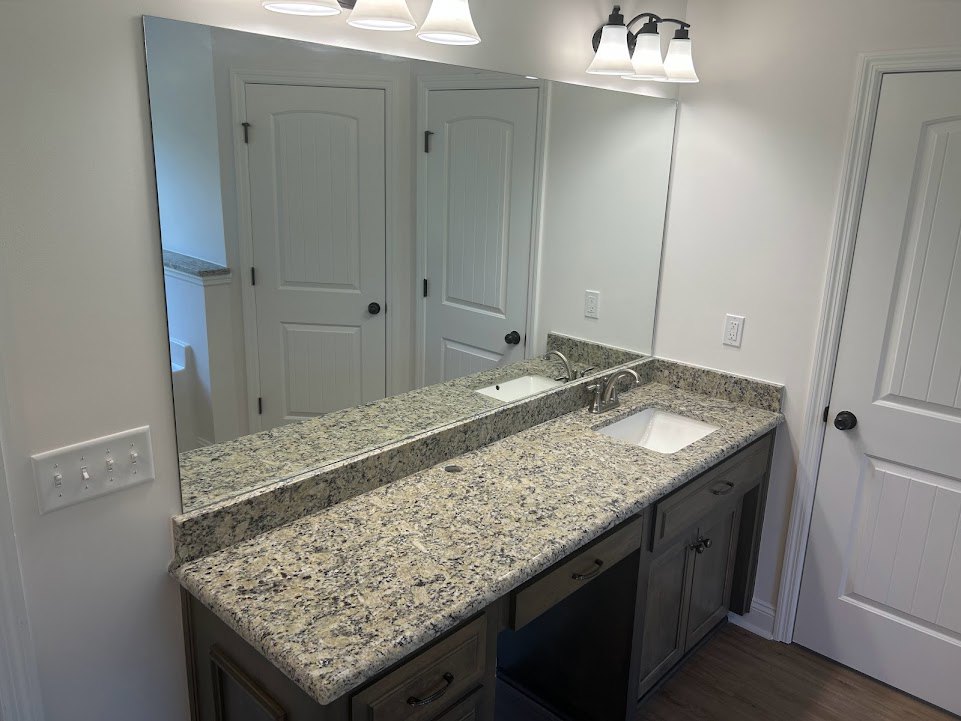 Bathroom with marble countertop, undermount white sink, large framed mirror, three-light fixture above, white cabinetry with black knobs, tile backsplash, and white door with black