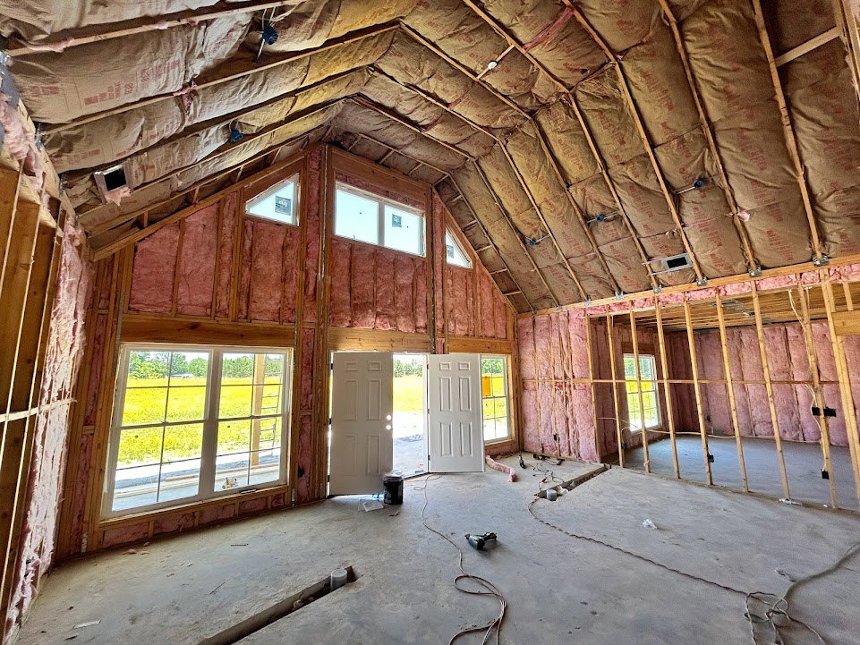 Exposed ceiling beams and unfinished walls in a room with a window overlooking a field, white door with cross detail, visible hole in the floor, signs of decay and building