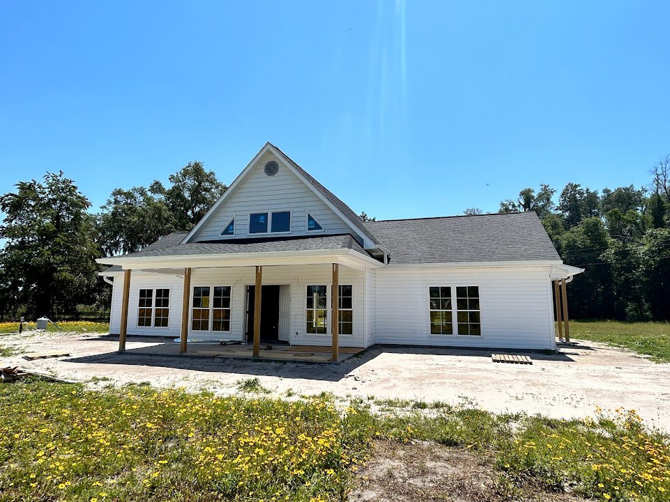 White siding house with wide covered porch, multiple windows with white trim, surrounded by green lawn and mature trees under blue sky