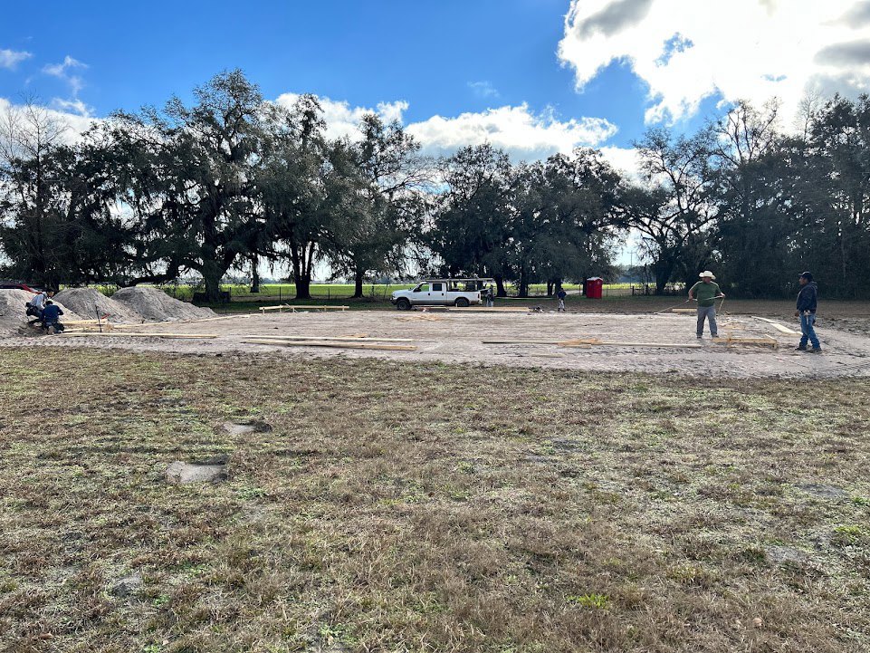 Man wearing a hat standing on bare dirt near a white truck, surrounded by grassy field and scattered trees under a partly cloudy blue sky