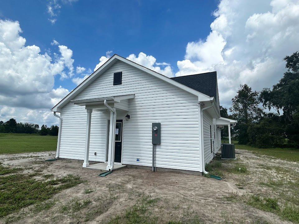 White siding house with black front door, black metal mailbox on stone post, dirt patch and grass in front yard, blue sky with scattered white clouds